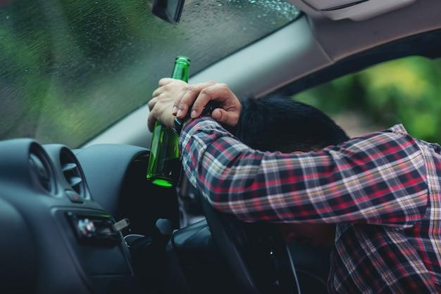 Person holding a green bottle, resting head on the steering wheel, symbolising drink driving